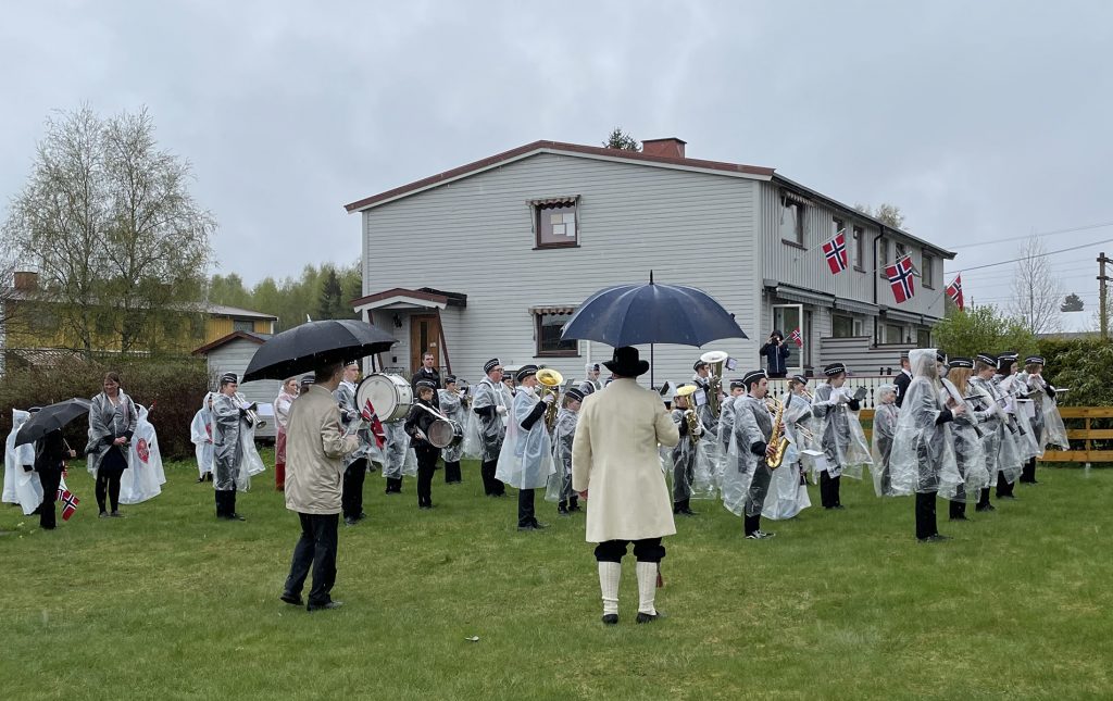 Jessheim skolekorps i regnponsjoer spiller marsj ved flaggstanga i Linjebo borettslag. 